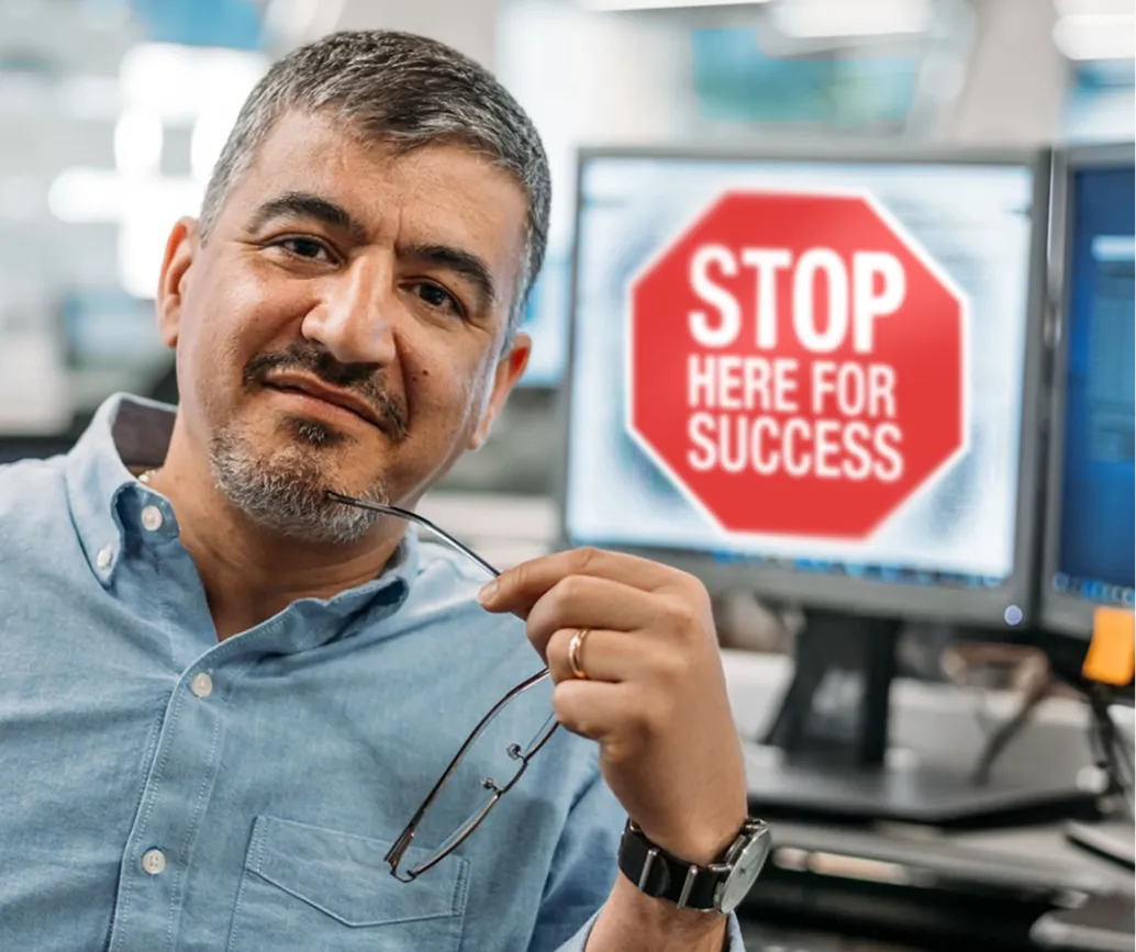 A corporate employee sitting in front of a computer with a sign on screen that says “Stop Here for Success.”