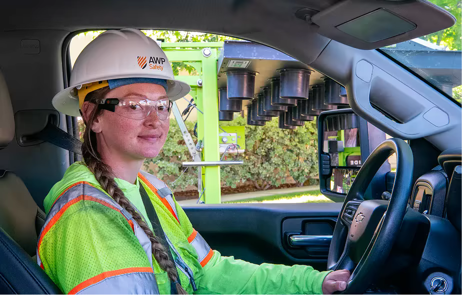 A woman wearing an AWP hardhat and visibility vest sitting in her work truck.