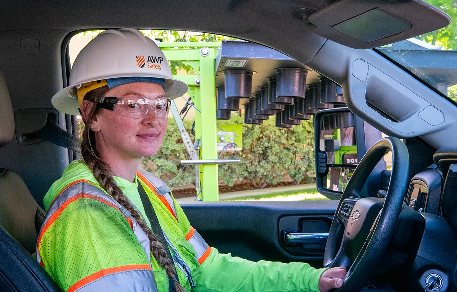 A woman wearing an AWP hardhat and visibility vest sitting in her work truck.
