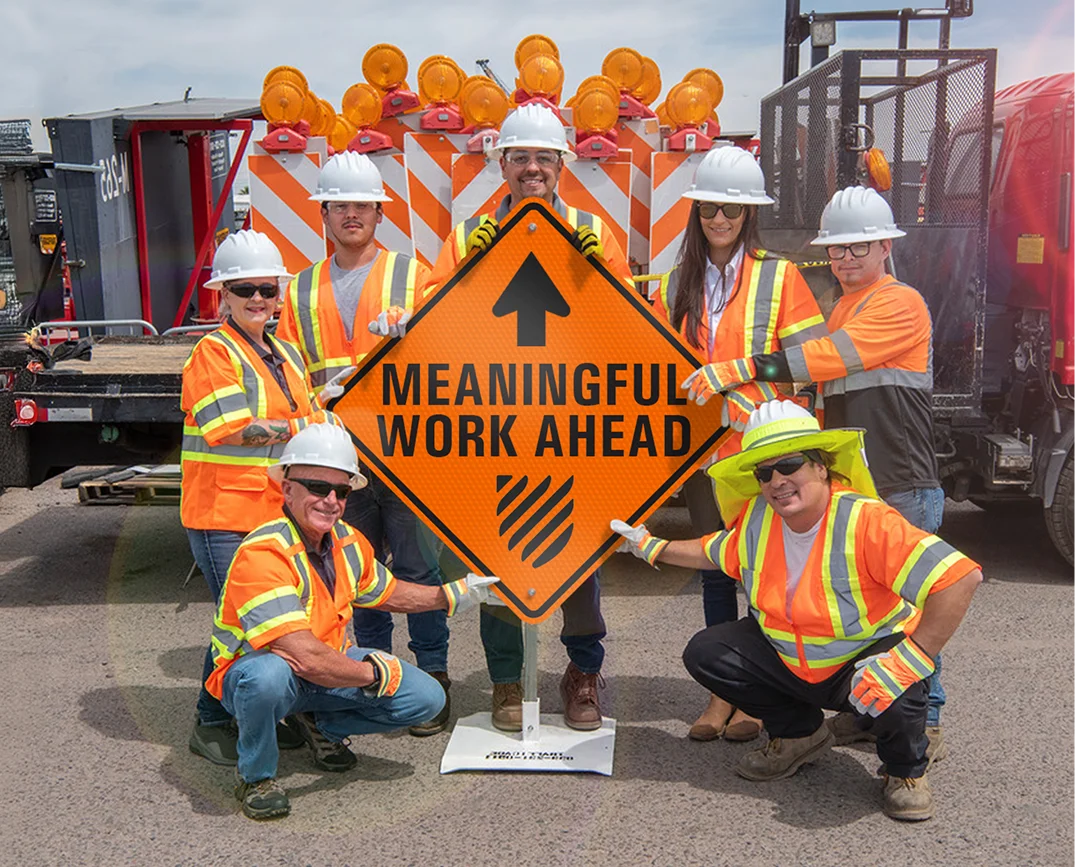 A group of AWP employees in orange visibility vests and hardhats pose with a traffic sign that says “Meaningful Work Ahead.”