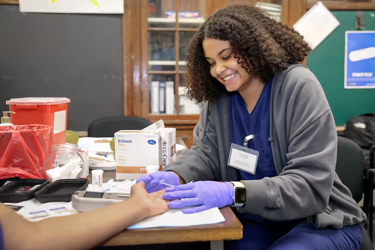 A Brown University Health professional performing a fingerstick blood test at a community clinic table with medical supplies.