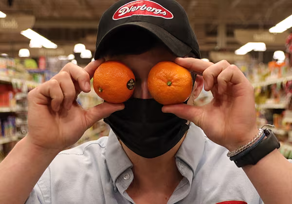 A produce associate holding two oranges up to his eyes and smiling
