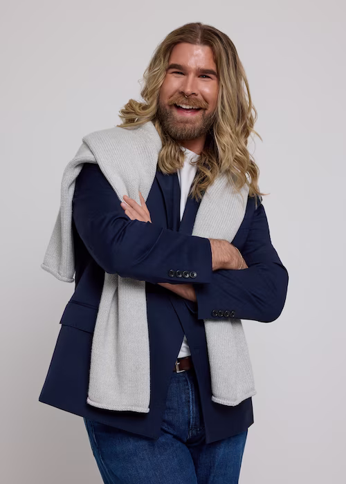 Stylish man with long wavy hair and beard, wearing a navy blazer, white shirt, and gray sweater draped over shoulders, standing confidently with arms crossed against a light background.