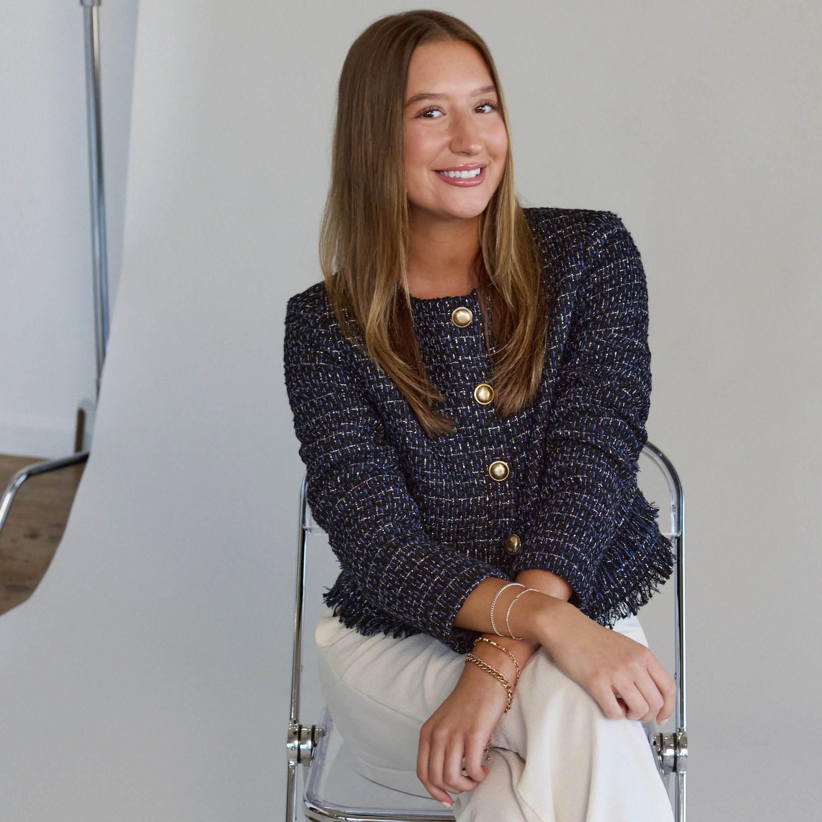 A person with long hair, wearing a textured jacket and light pants, sits on a clear chair smiling, with a neutral background.