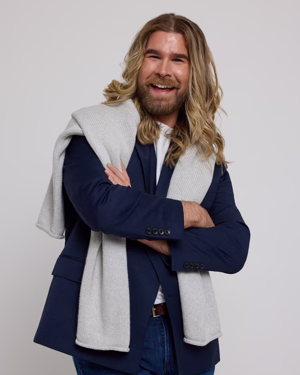 Stylish man with long wavy hair and beard, wearing a navy blazer, white shirt, and gray sweater draped over shoulders, standing confidently with arms crossed against a light background.
