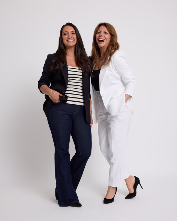 Two smiling women standing side by side, one in a black blazer, striped top and dark jeans, the other in a white suit and black heels.