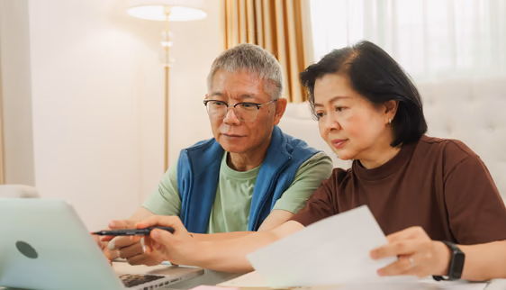 An older couple comparing paper forms to what they’re seeing on their laptop screen.