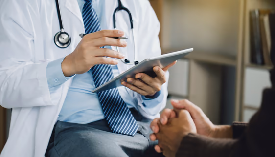 A doctor in a white coat using a tablet while speaking with a patient.