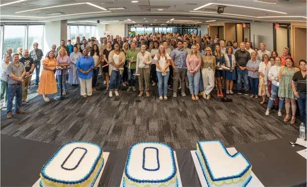 A group of people gathered at an event, celebrating Genesco's 100th anniversary with three cakes forming '100'.