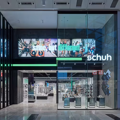 The front of a brightly lit Schuh store, one of the Genesco brands, along a city street.