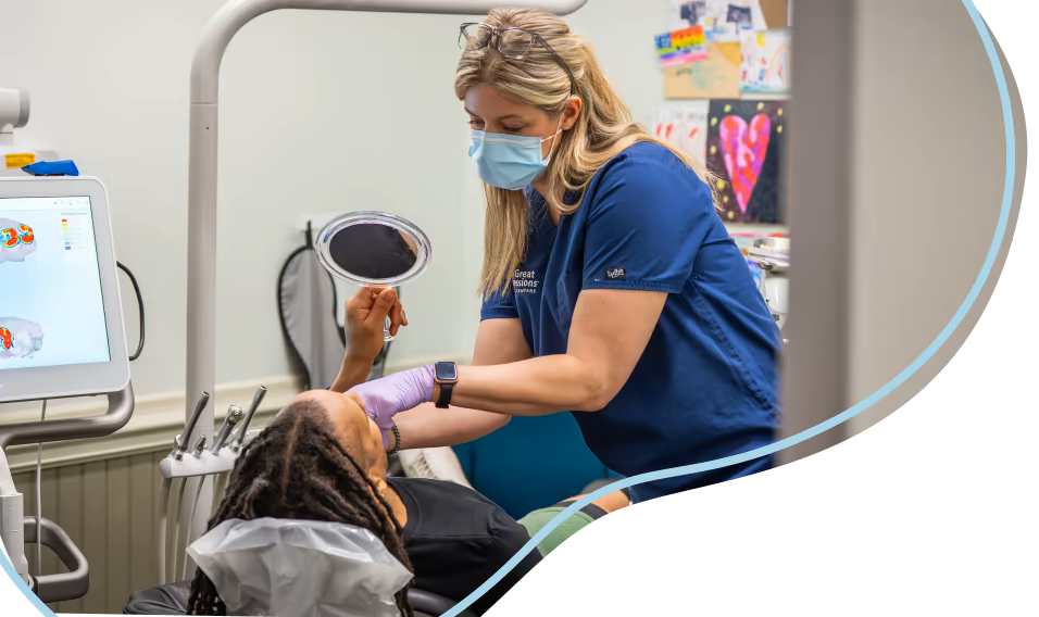 Dentist in blue scrubs and face mask with a pediatric patient looking in a mirror. 