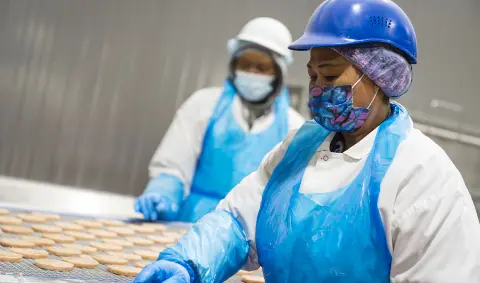 Two Koch Foods employees in hard hats, aprons, gloves and face masks inspecting food in a cooking facility