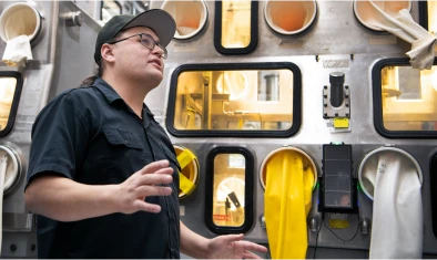 A person in a black shirt and cap stands in front of a metal lab machine with built-in gloves and clear windows, appearing to explain or demonstrate something
