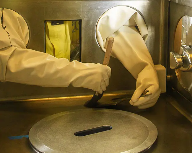 A pair of hands in protective rubber gloves reaching inside a glovebox chamber as part of an experiment.
