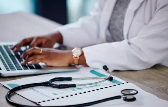 A healthcare professional typing on a laptop with a stethoscope laying on their desk.