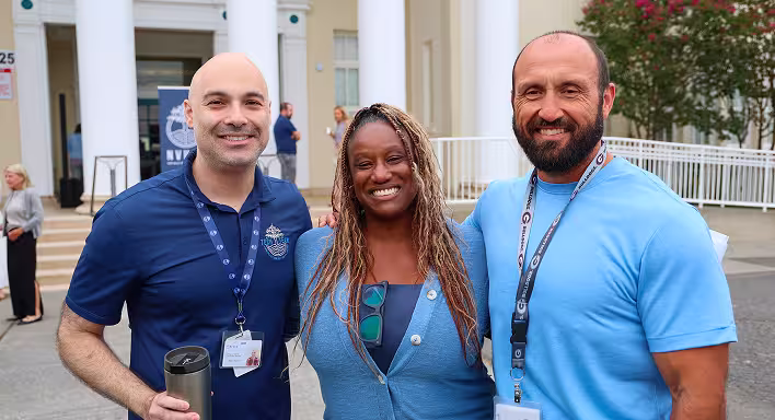 Three adults wearing blue shirts and ID lanyards standing in front of a building, smiling.