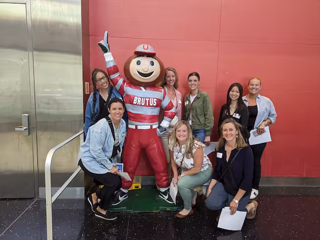 Eight staff members standing and kneeling around the Brutus Buckeye mascot for a group photo.