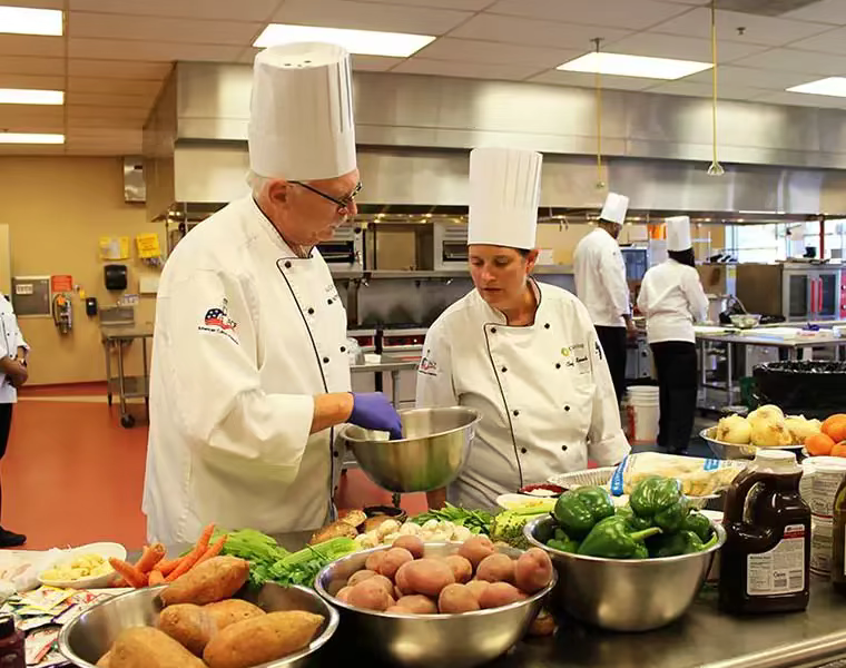 An older male chef consults with a female cook (both wearing white smocks and chef hats) in the kitchen of an Ohio Living facility.