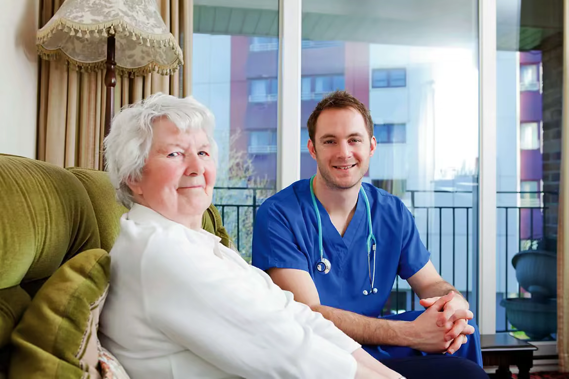 A smiling male CNA in blue scrubs sits and talks to a female resident in her room.
