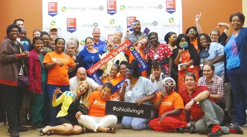A large group of smiling staff members stand or sit shoulder to shoulder in the lobby area of an Ohio Living facility.