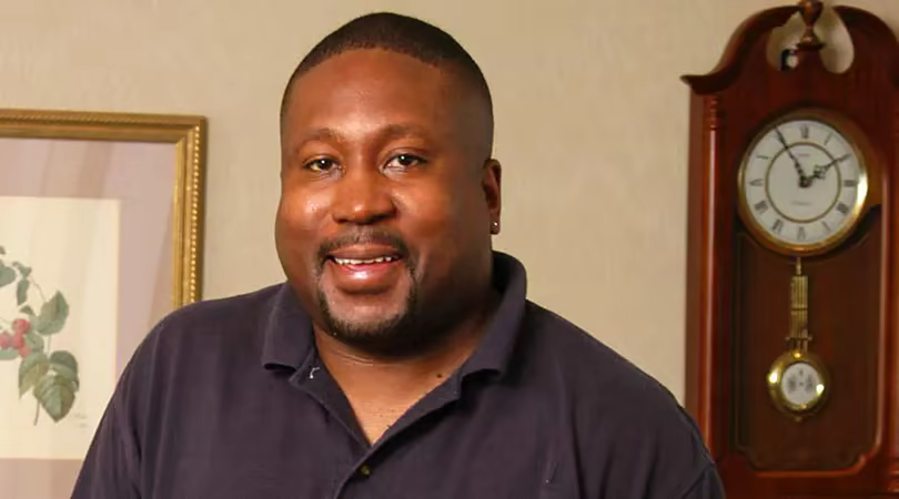 A Black, male staff member wearing a black polo shirt smiles as he stands next to a grandfather clock in a lobby.
