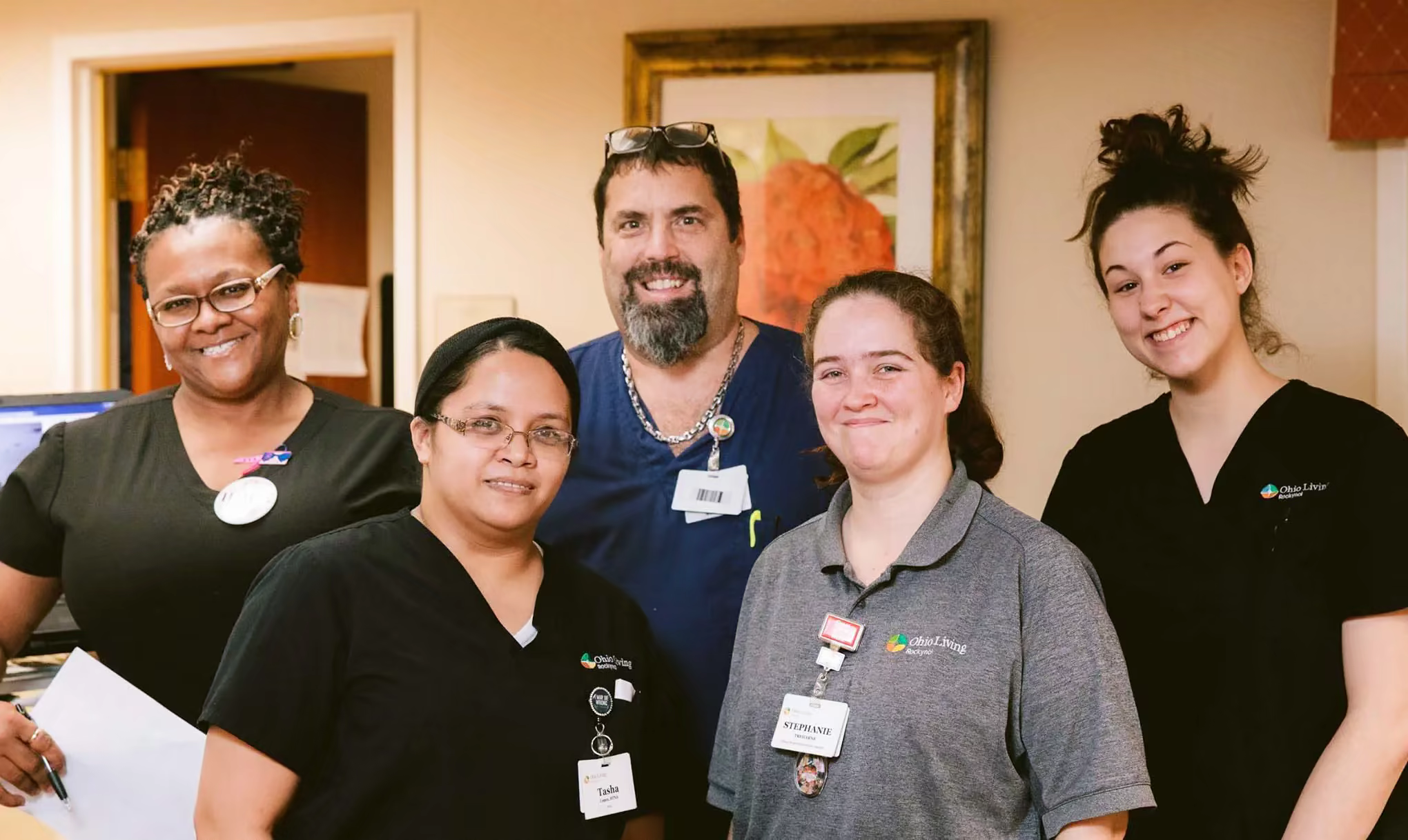 Four female and one male Ohio Living staff members in black scrubs pause to smile while they work at a station area.
