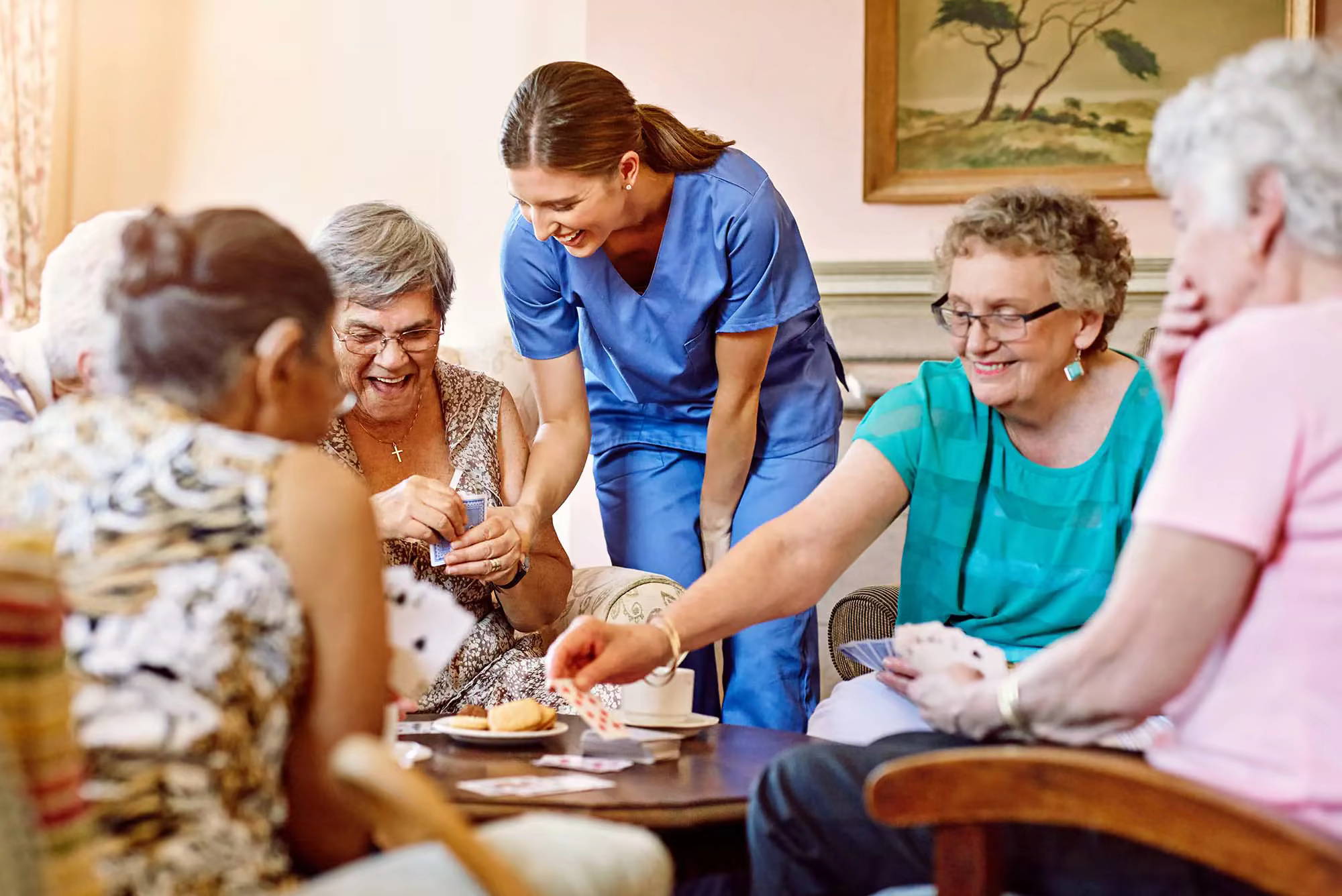 A female aide in blue scrubs assists four female residents who are playing cards.