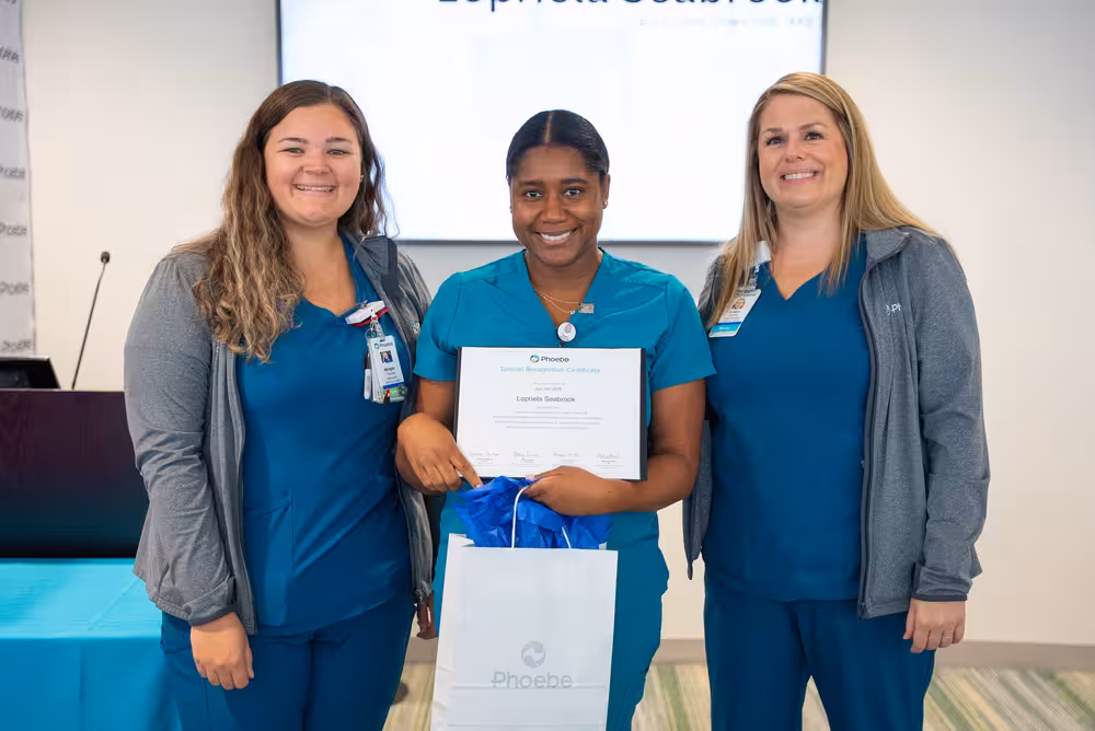 Two RNs standing on either side of a new RN holding an award, all smiling.