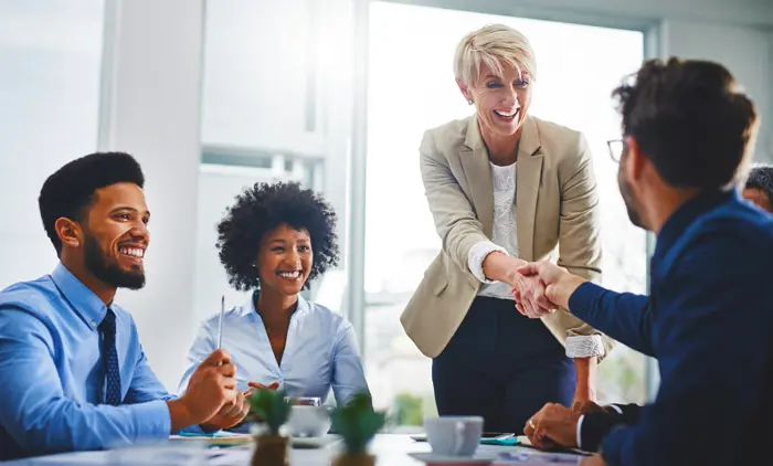 A businesswoman shaking hands with a person at a table while two other employees smile.