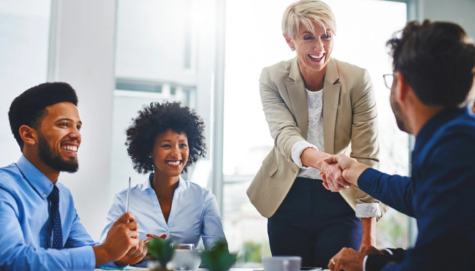 A businesswoman shaking hands with a person at a table while two other employees smile.