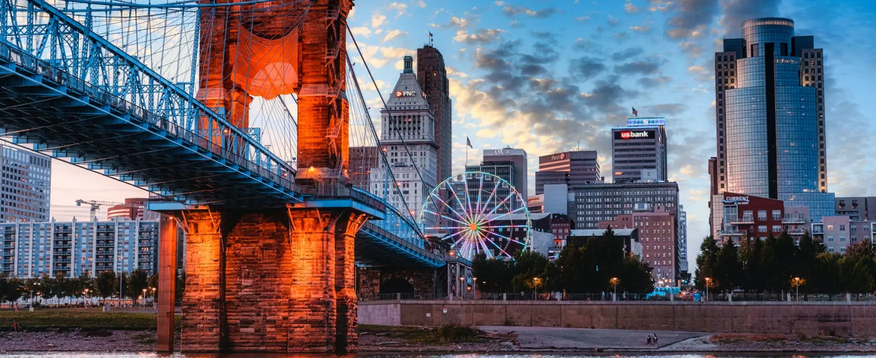  	The John A Roebling Suspension Bridge across the Ohio River and Cincinnati office buildings beneath a blue sky.