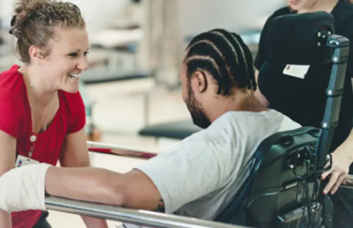 A smiling female therapist in red scrubs aide by a colleague works with a male patient in a wheelchair.