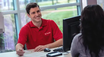 A young male customer service rep in a red polo assists a female client at his computer station.