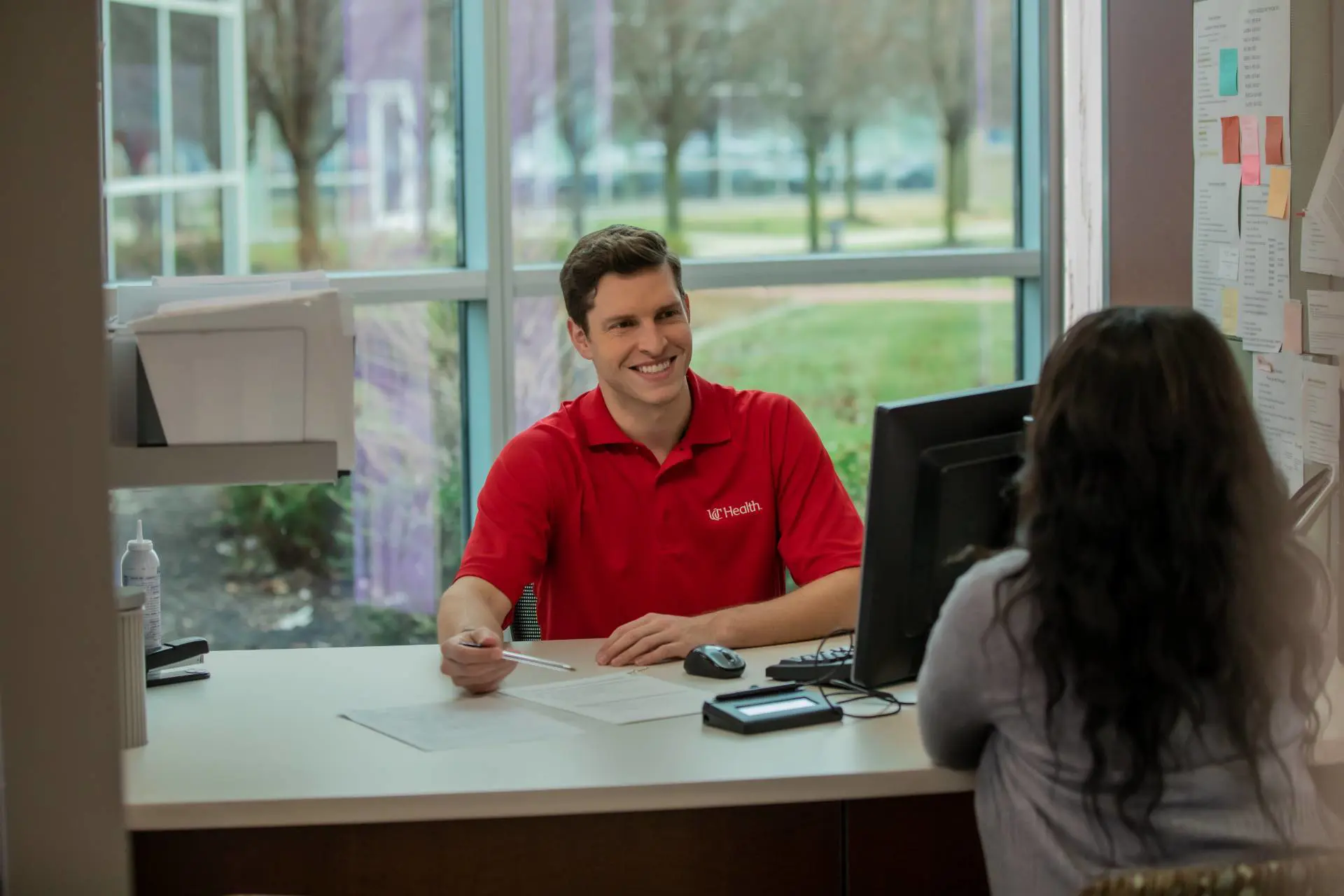  	A young male customer service rep in a red polo assists a female client at his computer station.