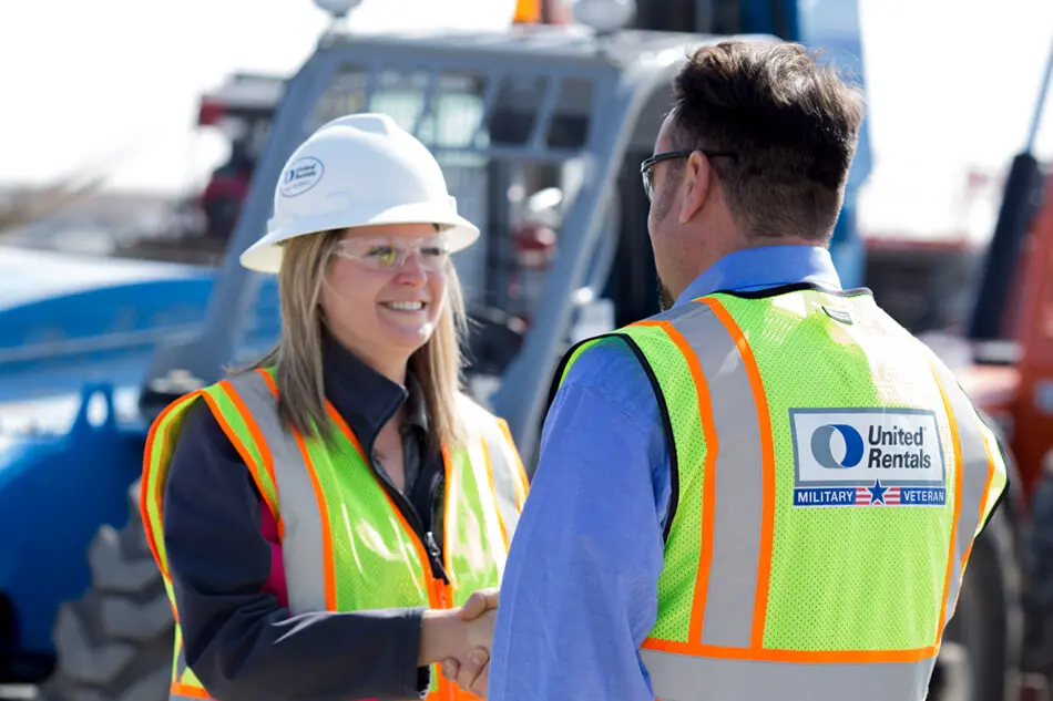 Une travailleuse souriante portant un casque et un gilet jaune s'entretient avec un collègue masculin sur un chantier de construction.