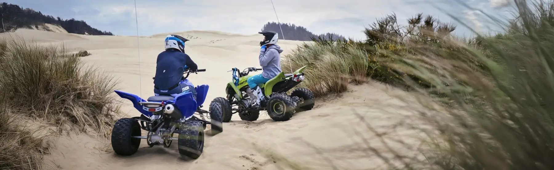 Two people riding Yamaha ATVs over sand dunes on a beach.