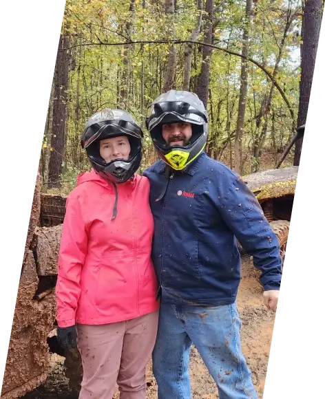 Two Yamaha employees in helmets pose next to a dirt bike track in a forest.