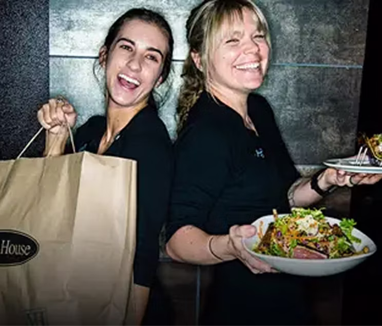 Two smiling to-go specialists holding a bag and two plates of food.