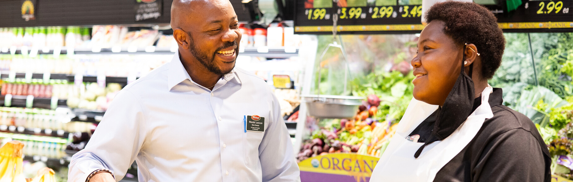 A smiling manager speaks to an employee in the produce section of a store.
