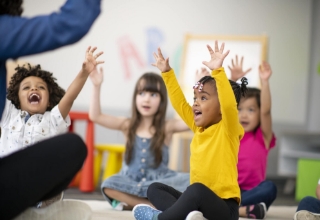 Children sitting down with their hands raised in excitement.