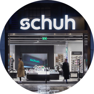 The front of a brightly lit Schuh store, one of the Genesco brands, along a city street.