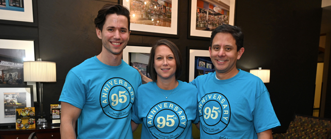 Three smiling Genesco employees in light blue t-shirts stand shoulder to shoulder in an office setting.