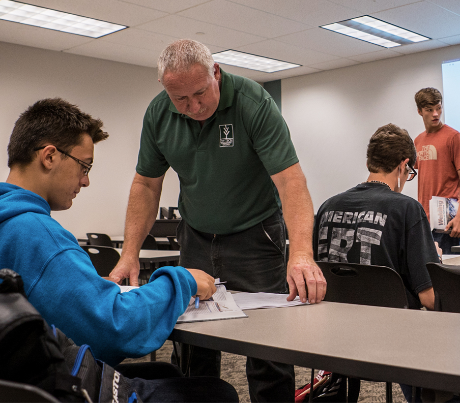 An older male faculty member conferences with a male student at the student&rsquo;s table in a classroom.