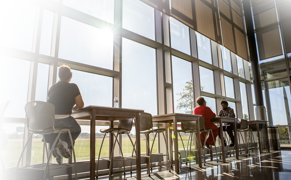 Three faculty members congregate and talk in a wide-open cafeteria/lounge area.