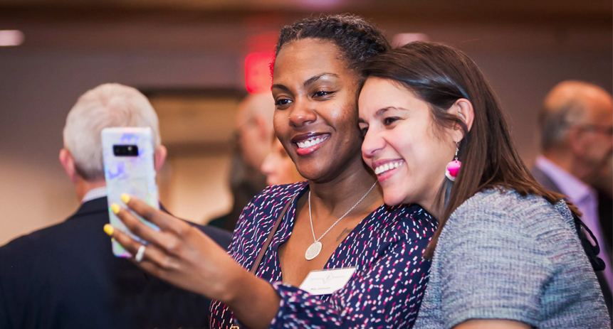 A white female faculty member takes a selfie with a Black female colleague during a break in a staff meeting.