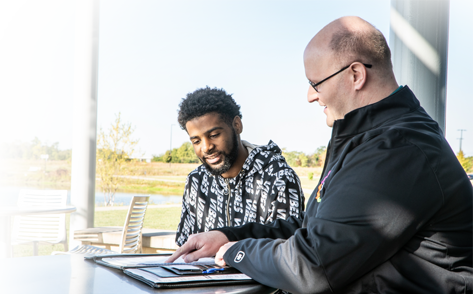 A white male faculty member conferences with a Black male student in a lounge area.