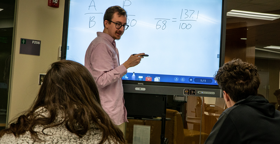 A male Ivy Tech faculty member points to the screen and lectures in the front of a classroom while students listen.