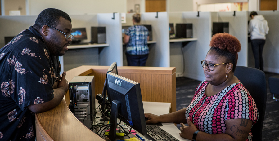 A Black male leans on a counter to talk to a Black female clerical employee who is gazing at her computer screen.