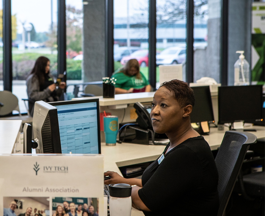 A Black female staff member sits at her desk in the Ivy Tech lobby and gazes at her computer screen.