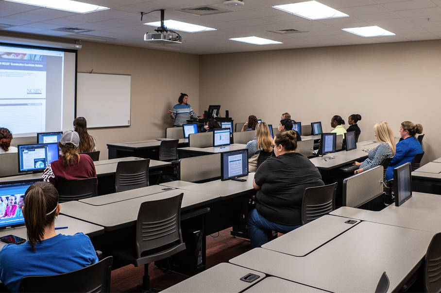 A group of students sit in a computer lab as a faculty member guides them through a lesson.